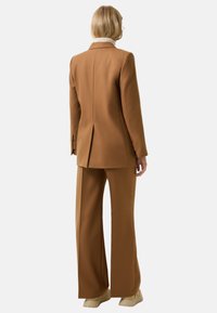 Osoba z krótkimi blond włosami, ubrana w brązowy skrojony garnitur i beżowe buty, stojąca tyłem na jednolitym tle.