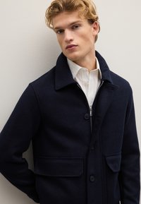 Cappotto in misto lana blu navy con chiusura con zip, frontale con bottoni, grandi tasche anteriori e colletto a rever abbinato a una camicia a righe bianche.