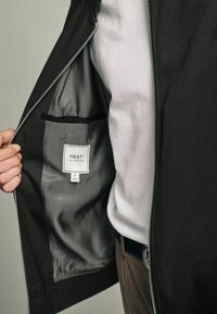 Crna jakna s patentnim zatvaračem i glatkom unutarnjom oblogom, s oznakom "NEXT Tailoring" koja sadrži upute za njegu i mali džep.