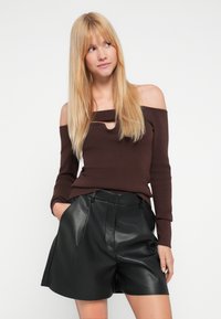 Bruine ribgebreide off-shoulder trui met een kleine opening bij de halslijn, gecombineerd met hooggetailleerde zwarte leren shorts met voorplooien.