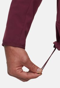 Main qui ajuste le cordon de serrage à la manche d'un manteau ou chemise à manches longues maroon avec un système de fermeture élastique.