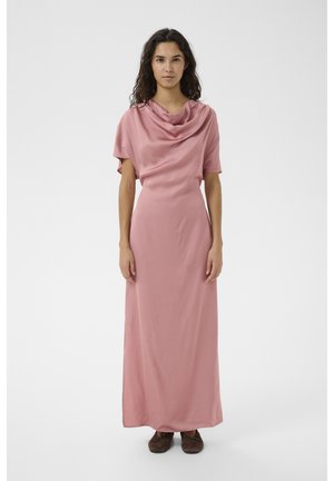 Kobieta o długich ciemnych włosach ubrana w różową suknię sięgającą podłogi z dekoltem w kształcie drapowanego materiału i brązowe buty, stojąca na jednolitym tle.