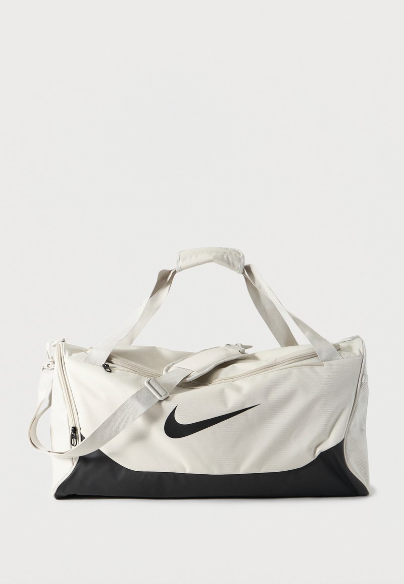 Λευκή και μαύρη τσάντα τύπου duffel της Nike με ρυθμιζόμενο ιμάντα ώμου και διπλές λαβές, εμφανίζεται πάνω σε απλό φωτεινό φόντο.