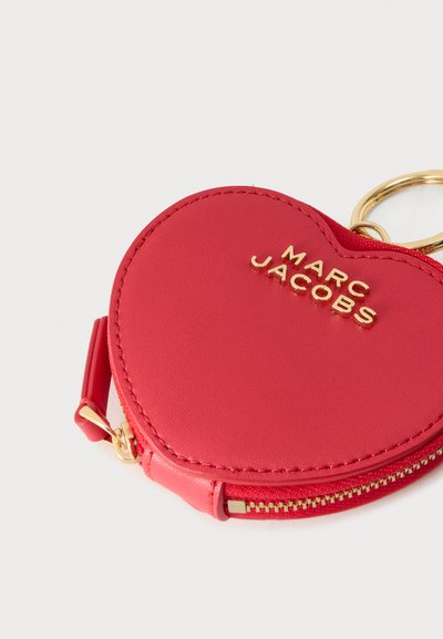 Κόκκινο δερμάτινο πορτοφολάκι σε σχήμα καρδιάς με χρυσλό φερμουάρ, χρυσό μπρελόκ κλειδιών και το όνομα «MARC JACOBS» ανάγλυφο σε χρυσά γράμματα μπροστά.