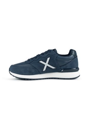 Zapatilla deportiva azul oscuro con el logo blanco "X" en el lateral, cordones negros y suela blanca con dibujo negro, mostrada desde el lado izquierdo sobre fondo blanco.