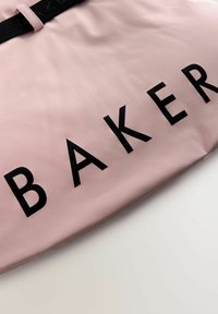 Rozā auduma soma ar uzrakstu „BAKER” lieliem melniem burtiem, ar gludu tekstūru un saskaņotu melnu siksnu.