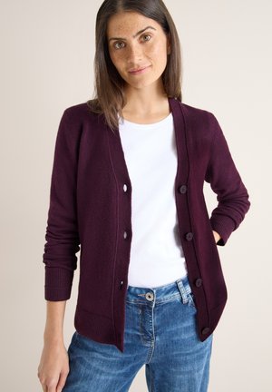 Bordeaux cardigan gemaakt van zachte stof; heeft een V-hals, lange mouwen en vijf knopen aan de voorkant; gecombineerd met een wit shirt en blauwe jeans.