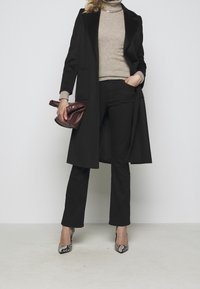 Schwarzer knielanger Mantel mit Reverskragen, beiger Rollkragenpullover, schwarze Hose und high heels im Schlangenmuster. Trägt eine burgunderrote Handtasche.