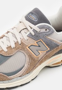 Harmaa ja beige tennari, jossa on verkko- ja mokkanahkaseoksia, oranssi yksityiskohta sekä teksturoidut yksityiskohdat logon ja nauhojen ympärillä.