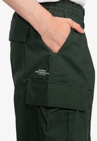 Zelené cargo nohavice vyrobené z textúrovaného materiálu, s elastickým pásom, bočnými vreckami a bielym vyšitým logom.