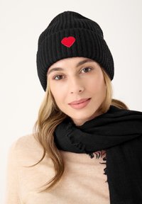 Zwarte geribde gebreide beanie met een rode hartborduring aan de voorkant. Het model draagt een zwarte sjaal, gestyld met een lichtbeige sweater.