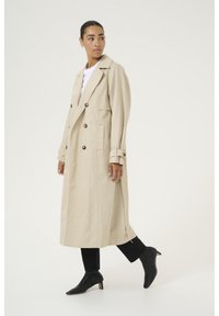 Bež trench kaput s dvostrukim gumbima, izrezanim ovratnikom, dugim rukavima, bočnim prorezima i slobodnim krojem, uparen s crnim čizmama s visokim potpeticama.