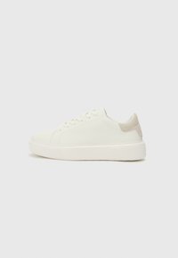 Baskets basses blanches avec une texture lisse, présentant un accent en daim beige clair sur le talon et une épaisse semelle en caoutchouc blanche.