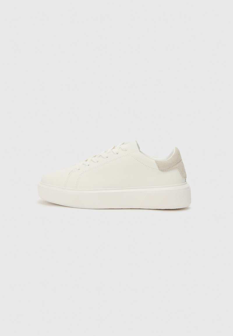 Baskets basses blanches avec une texture lisse, présentant un accent en daim beige clair sur le talon et une épaisse semelle en caoutchouc blanche.