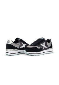 Zapatillas deportivas en negro y gris con parte superior de ante y malla, acentos blancos, suela estampada y un destacado logo en forma de X a los lados.