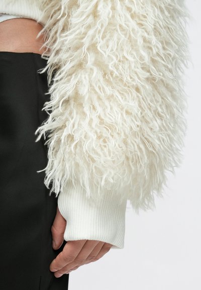 Κοντινό πλάνο ενός ατόμου που φοράει λευκό μπουφάν με υφή αφράτης γούνας και ριμπ μανσέτες, με μαύρο παντελόνι, το χέρι χαλαρά δίπλα στο σώμα.