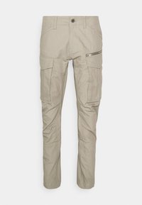 Pantalon cargo beige avec plusieurs poches, y compris des poches à rabat et zippées, passants de ceinture et coutures renforcées au niveau des genoux.