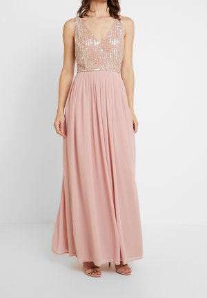 Vrouw draagt een mouwloze blush roze avondjurk met een paillettenversierde V-hals top en een vloeiende geplooide rok, gecombineerd met nude hakken.