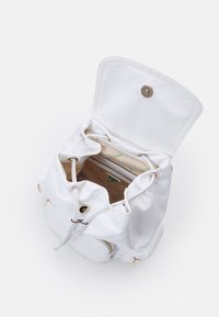 Mochila de tela blanca con cierre de cordón, herrajes en tono dorado y un interior beige con un patrón de logo verde y un bolsillo con cremallera.