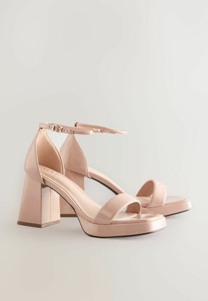 Roza lakirana usnjena sandala na visoki peti z blok peto, odprtim prstom in nastavljivim gležnjevim trakom, ki ima gladek, sijoč zaključek.