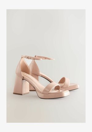 Roza lakirana usnjena sandala na visoki peti z blok peto, odprtim prstom in nastavljivim gležnjevim trakom, ki ima gladek, sijoč zaključek.