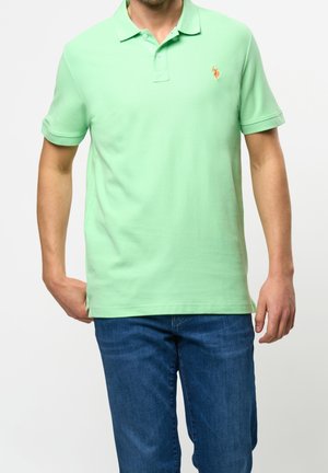 Homme portant un polo vert clair à manches courtes avec un logo brodé orange sur la poitrine gauche et un jean bleu sur fond blanc.
