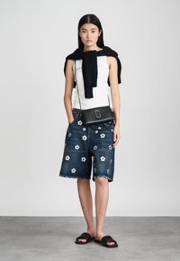 Denimshorts med hvit blomsterbroderi, en hvit tanktopp og en svart strikket genser knyttet rundt skuldrene; kombinert med svarte slides.