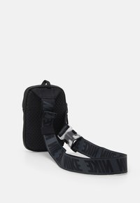 Liten svart texturerad påse med bred svart rem som har "NIKE"-text och en silverfärgad spänne, placerad mot en vit bakgrund.