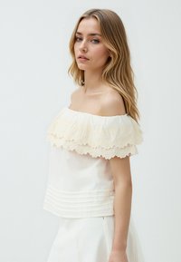 Młoda kobieta z długimi blond włosami, ubrana w białą bluzkę z odkrytymi ramionami z koronkowymi detalami oraz białą spódnicę, stojąca nieco bokiem.