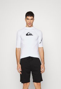 White short-sleeve rash guard with a black logo on the chest and sleeve, paired with black drawstring swim shorts. Smooth texture, fitted design.