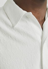 Getextureerde witte blouse met een subtiel bloemenpatroon. Heeft een puntige kraag en een knoopje bij de hals. De stof lijkt zacht en lichtgewicht.