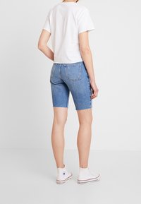 Modré džínové šortky s třepícím se lemem, doplněné bílým bavlněným tričkem a bílými vysokými teniskami; pohled zezadu.