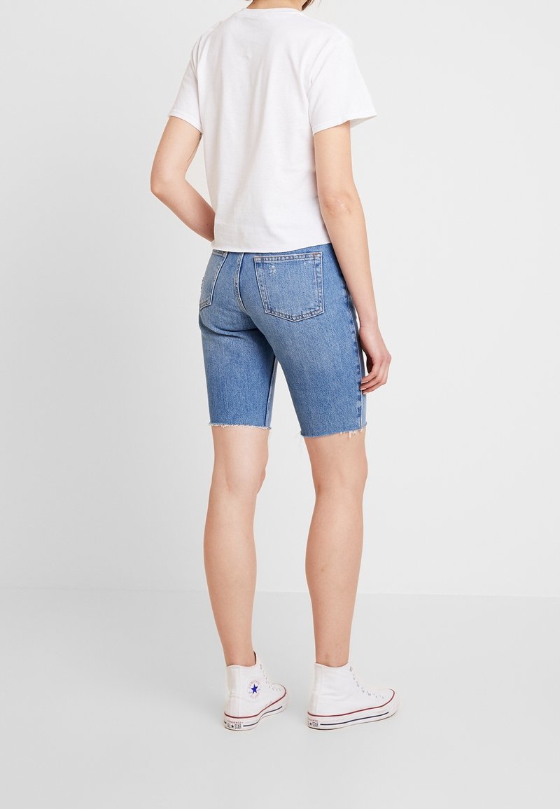 Modré džínové šortky s třepícím se lemem, doplněné bílým bavlněným tričkem a bílými vysokými teniskami; pohled zezadu.