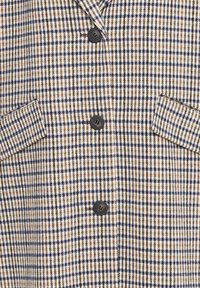 Primo piano di un tessuto a scacchi beige, blu navy e bianco con tre bottoni neri e due tasche a pattina inclinata.