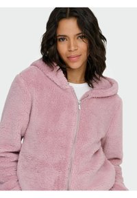 Różowa, miękka bluza z kapturem na zamek, wykonana z delikatnej syntetycznej tkaniny o gładkiej fakturze i z przodu zapinana na zamek.