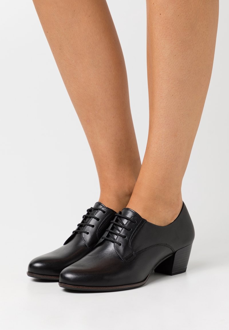 Chaussures montantes en cuir noir avec talons bas empilés, portées pieds nus sur fond blanc.