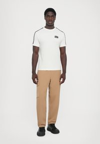 White short-sleeve t-shirt with black side accents and a chest logo, paired with light brown cargo pants and black sneakers.