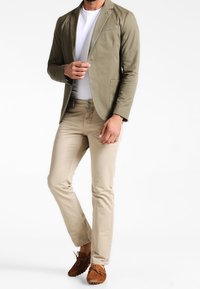 Oliwkowo-zielona marynarka, biała koszulka, beżowe spodnie i brązowe mokasyny. Marynarka ma klapy z wcięciem i jest dopasowana. Spodnie mają prosty krój.