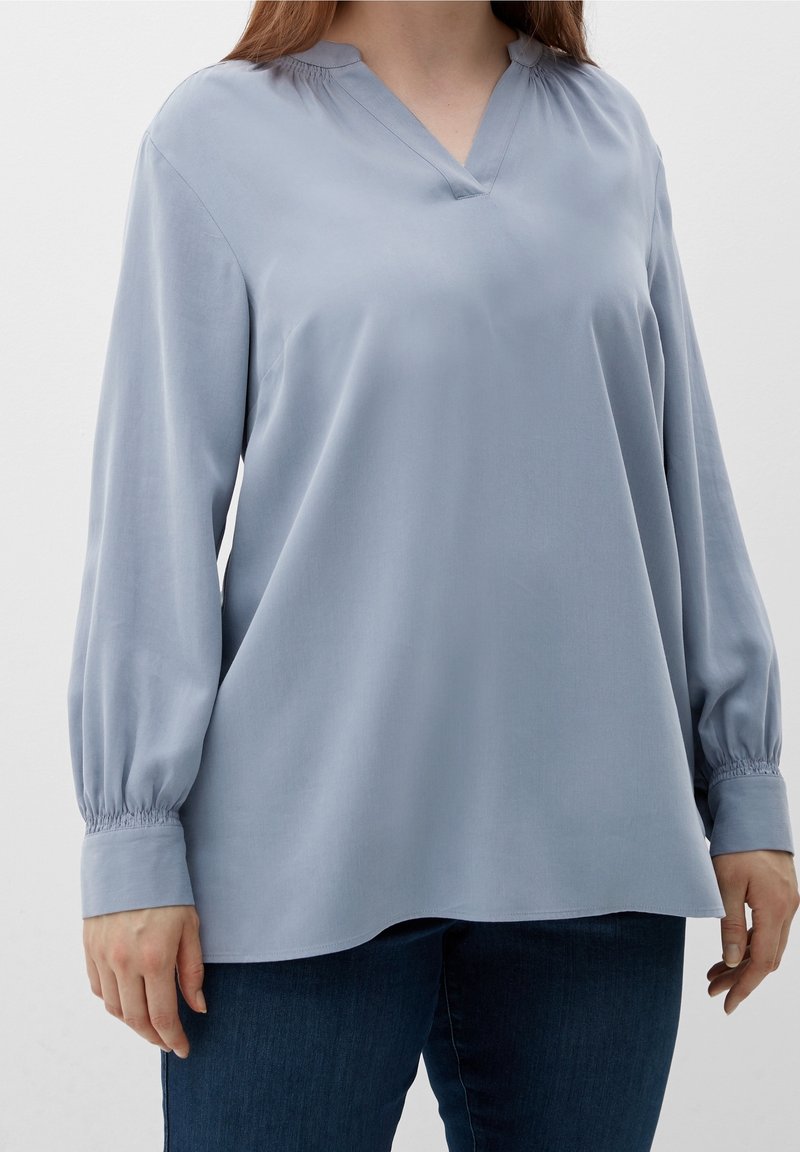 Helle blaue Bluse mit langen Ärmeln, V-Ausschnitt und gerafften Manschetten. Glattes Material mit lockerer Passform, leicht abgerundetem Saum und minimalistischem Design.