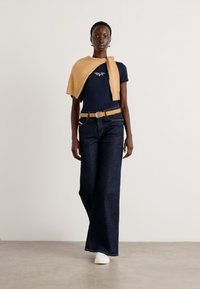Marineblå t-skjorte, lysebrun genser lagt over, mørkeblå vide jeans og hvite joggesko. Belte med brune detaljer. Enkel, moderne design.