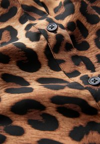 Tyg med leopardmönster i tan och svart, med en knappdetalj. Texturen verkar slät och något glansig.