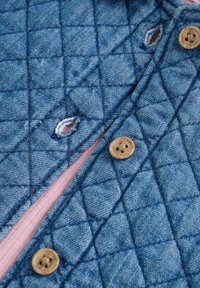 Gequilte spijkerjas met een lichtblauwe buitenkant, voorzien van bruin knopen en een roze voering. Versleten accenten nabij de knoopsgaten.