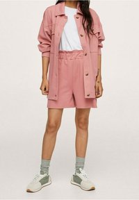 Femme portant une veste rose à boutons avec des manches retroussées, un short rose assorti, un t-shirt blanc, des chaussettes grises et des baskets blanches avec des détails verts.