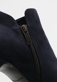 Navy semišové kotníkové boty se zipem na boku, zaoblenou špičkou a hladkým černým podpatkem. Obsahují jemné švy pro přidaný detail.