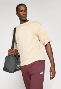 Beige tričká s krátkym rukávom, vo voľnom strihu, skombinované s bordovými teplákmi s malým modrým logom. Čierna športová taška s logom držaná v ruke.