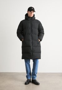 Black puffer coat with a hood, zip closure, and side pockets. Worn over light blue jeans and black boots, set against a neutral background.