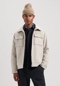 Light gray wool jacket with two front pockets, standing collar; worn over a dark shirt and paired with a beige knit beanie.