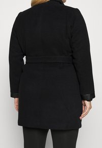 Manteau en laine noir avec une taille ceinturée, un col à crans et des poignets en cuir. Texture lisse et design classique, adapté aux temps plus froids.