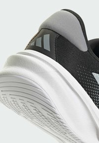 Baskets de sport noires avec une tige texturée, des accents gris et une semelle en caoutchouc blanche dotée de rainures pour une meilleure traction. Détail du logo visible.