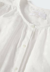 Blouse blanche avec de petites fleurs brodées, col rond, fermeture boutonnée devant et détail froncé près du col.